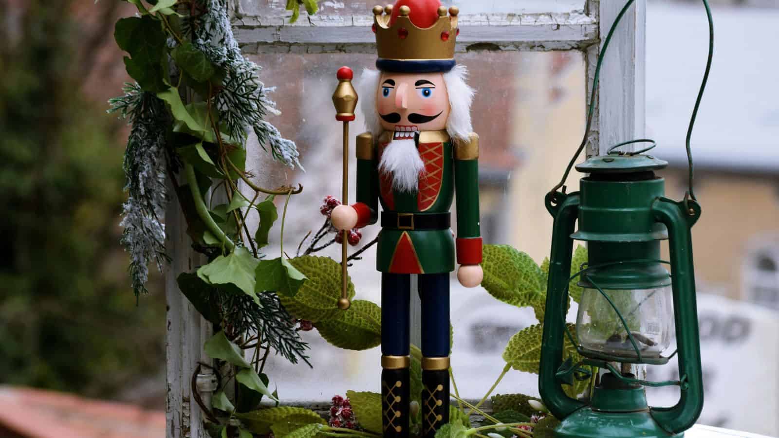 A nutcracker figurine dressed as a king stands on a windowsill, surrounded by green foliage and a vintage lantern. The window offers a glimpse of an outdoor scene with blurred buildings in the background.