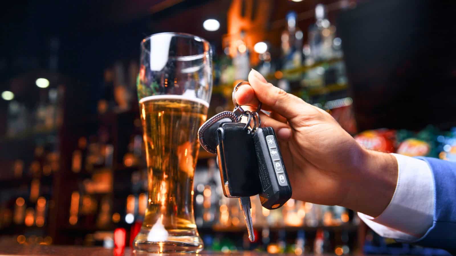 A hand is holding a set of car keys in front of a tall glass of beer on a bar counter. The background is blurred with various bottles on shelves.