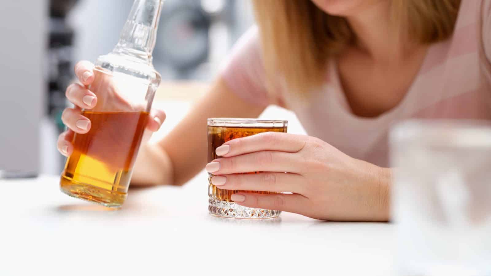 A person pours amber-colored liquid from a bottle into a glass on a white countertop. The person's nails are painted light pink. The scene suggests a casual setting with a focus on the drink preparation.