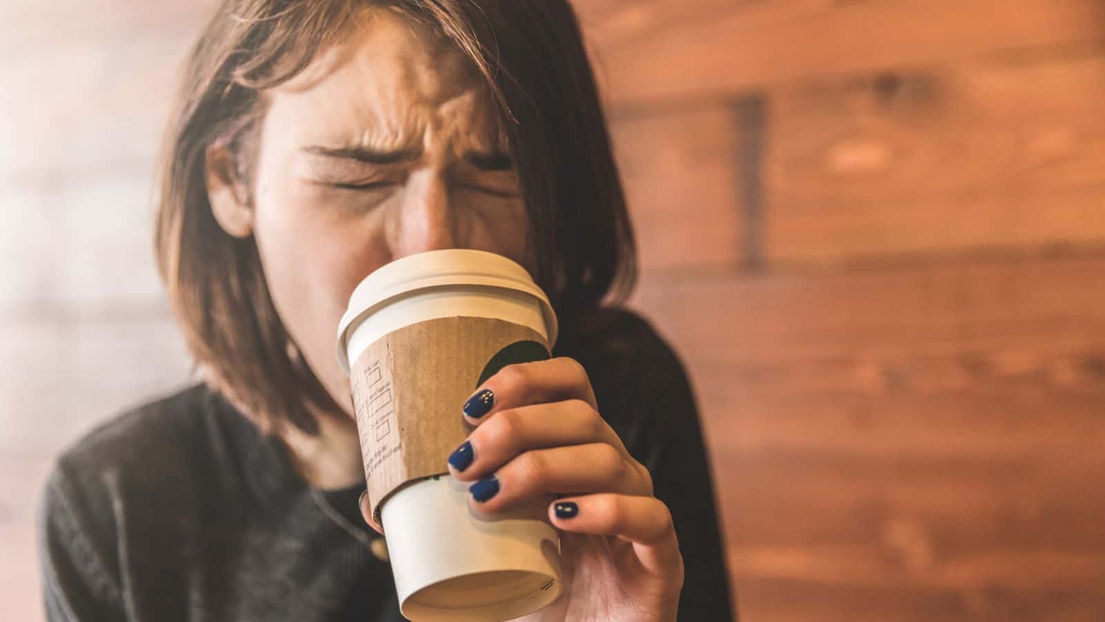 A person with short hair and dark nail polish is holding a takeaway coffee cup close to their face with a pained expression. The background is a wooden wall.