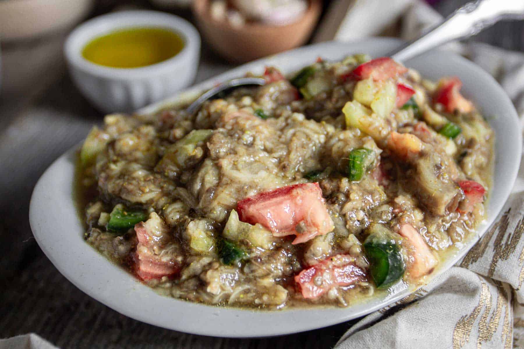 A white plate holds a dish of mashed eggplant mixed with diced tomatoes and green peppers. A small bowl of olive oil is visible in the background, along with a cloth napkin and a metal spoon partially submerged in the eggplant mixture.