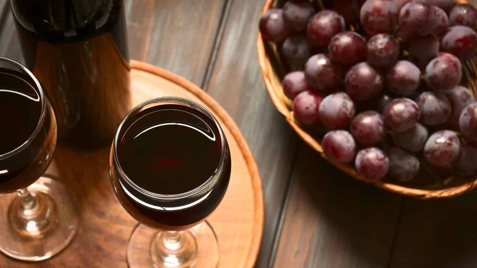 Two glasses of red wine sit on a wooden tray beside a basket of purple grapes. A wine bottle is partially visible in the background on a wooden surface.