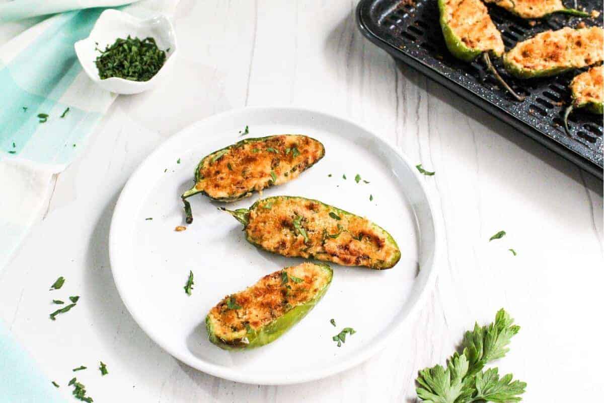 A white plate with three baked jalape&ntilde;o poppers, topped with herbs, rests on a white surface. In the background, a baking tray teems with more showstopping appetizers and a small bowl of chopped herbs. Parsley is scattered artfully on the table.