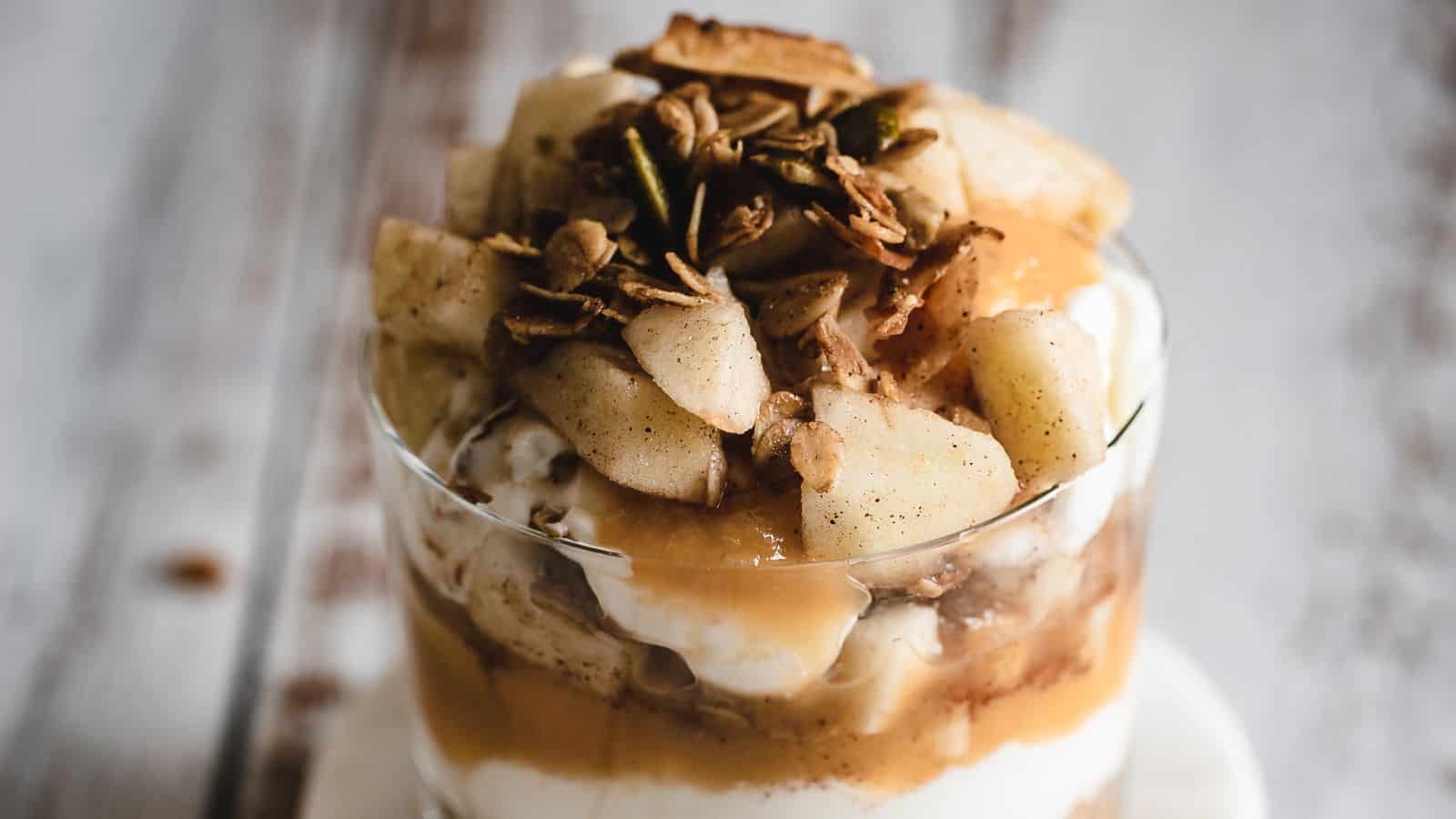 A glass parfait with layers of yogurt, granola, sliced apples, and a drizzle of caramel sauce, topped with additional granola and apple pieces. It is placed on a light-colored wooden table.