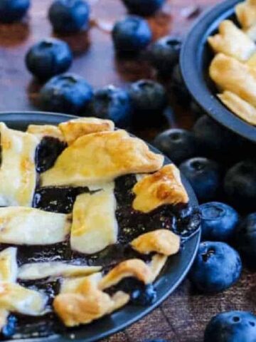 Two small blueberry pies with golden lattice crusts on a wooden table, surrounded by scattered fresh blueberries. A silver fork rests nearby.