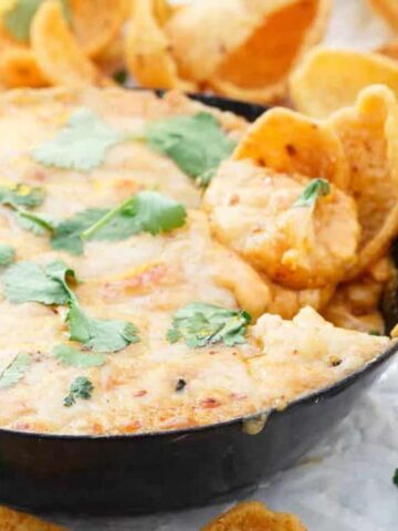 A skillet filled with melted cheese topped with cilantro. Surrounding the skillet are corn chips, some dipped in the cheese. A lime and more cilantro are visible in the background.