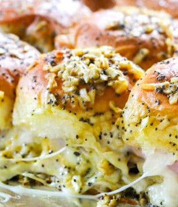 A tray of freshly baked slider sandwiches with glossy tops, sprinkled with minced garlic and herbs. The sandwiches are filled with melted cheese and thin slices of ham, with cheese oozing out, sitting on a lined baking tray.