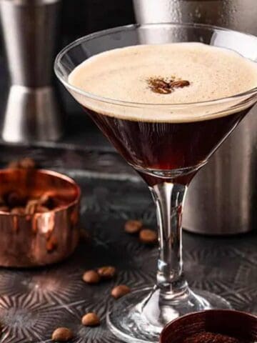 A martini glass filled with an espresso martini sits on a textured tray. Coffee beans and a scoop are on the tray. A stainless steel cocktail shaker and jigger are in the background.