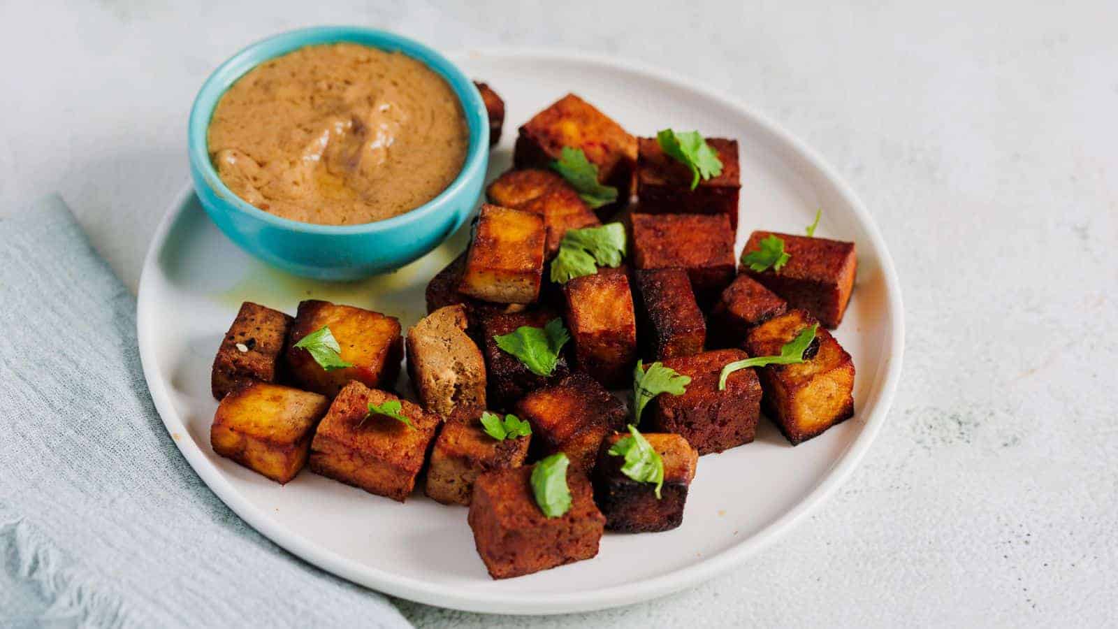 A white plate with crispy tofu cubes garnished with cilantro. A small blue bowl of creamy brown dipping sauce is placed on the plate. The plate is on a light gray surface with a folded gray cloth nearby.