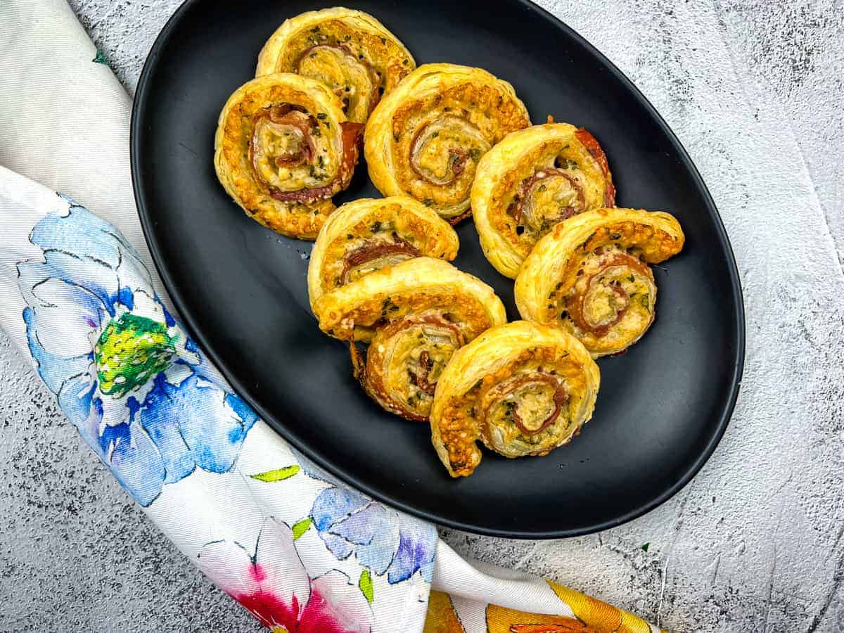 A plate of spiral puff pastry bites filled with meat and cheese is placed on a black oval dish. The dish is on a textured gray surface, partially resting on a colorful floral cloth.