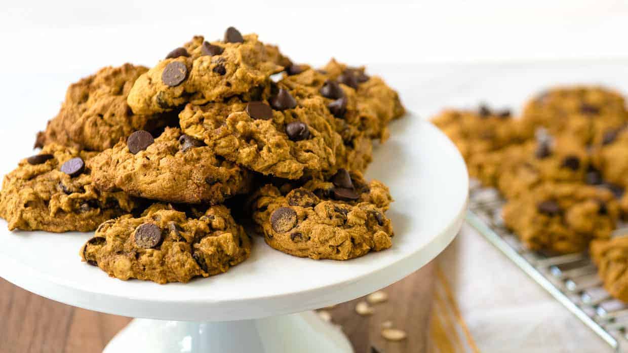 Pile of pumpkin oatmeal chocolate chip cookies with more in the background on a cooling rack.