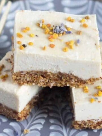 A plate with three stacked pieces of creamy dessert bars, topped with small yellow and purple sprinkles. The bars have a crumbly brown base. A fork is placed next to the bars on the patterned plate.