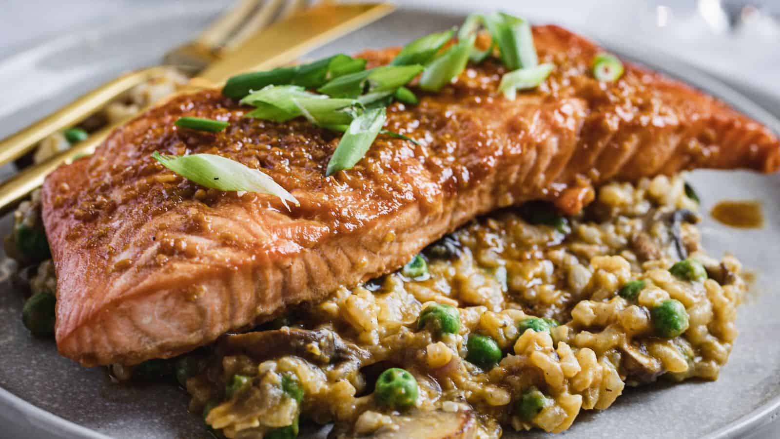 A plate of roasted salmon topped with sliced green onions is served over a bed of creamy risotto with peas and mushrooms. A fork and knife are placed beside the dish.
