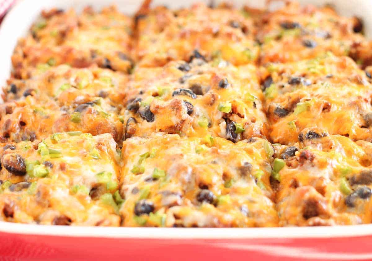A close-up of a cheese-laden casserole with visible olives, green peppers, and other toppings, cut into square portions and nestled in a red baking dish.