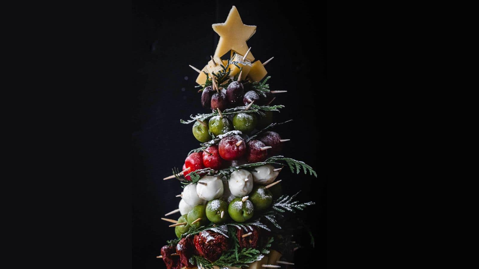 A festive tree-shaped arrangement made of assorted vegetables and cheeses is topped with a star-shaped cheese. It includes olives, cherry tomatoes, bocconcini, and is decorated with sprigs on a dark background.