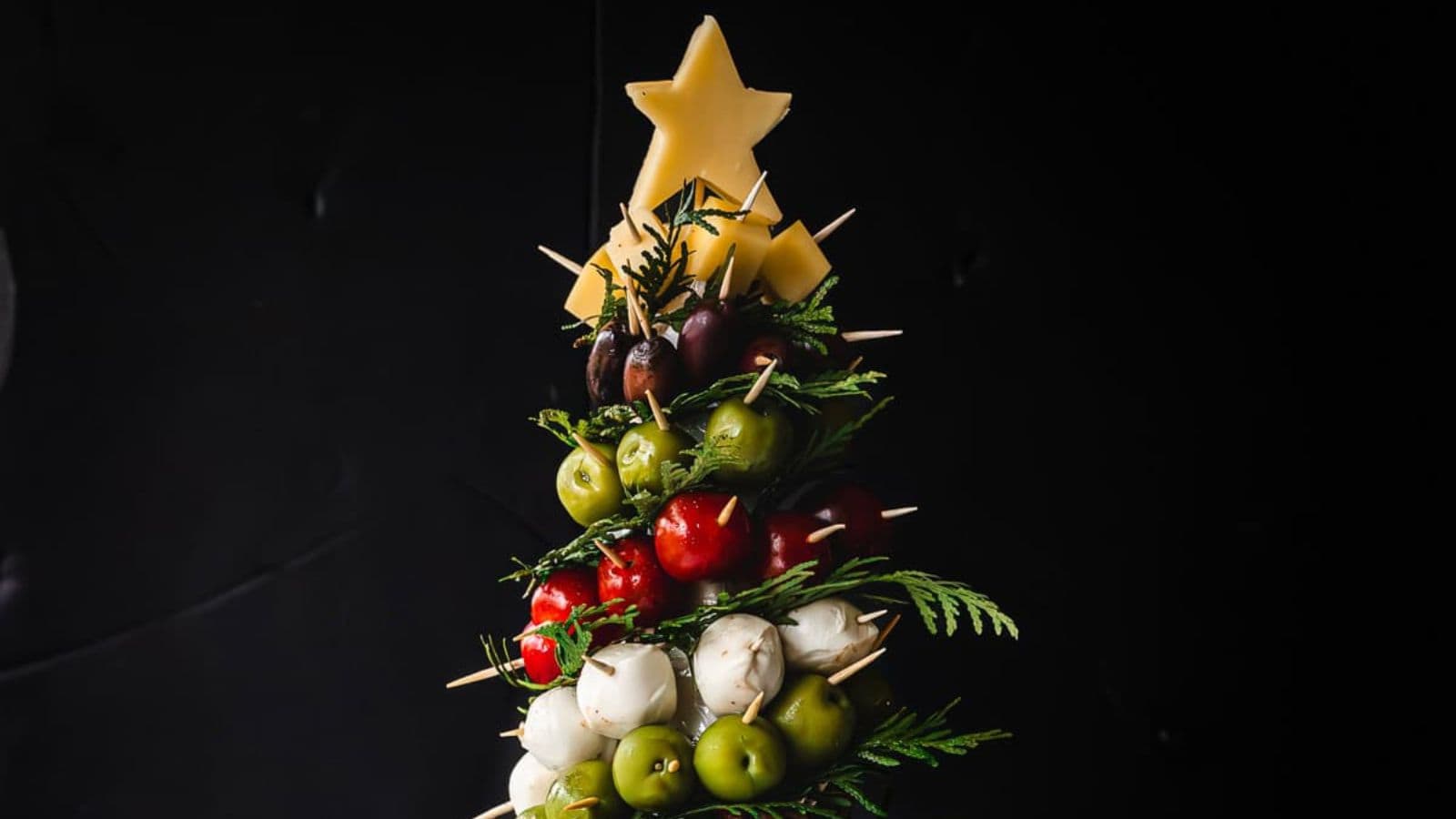 A festive holiday skewer arranged in the shape of a Christmas tree with layered tomatoes, mozzarella balls, olives, and herbs. The top is adorned with star-shaped cheese pieces. The background is dark, highlighting the vibrant colors of the arrangement.