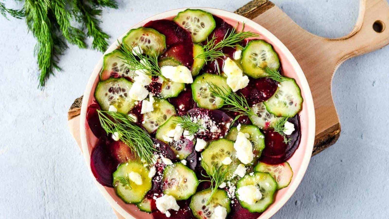 A bowl of salad containing sliced cucumbers and beets, topped with crumbled feta cheese and fresh dill. The bowl is placed on a wooden cutting board, and a sprig of dill is lying beside it on a light-colored surface.