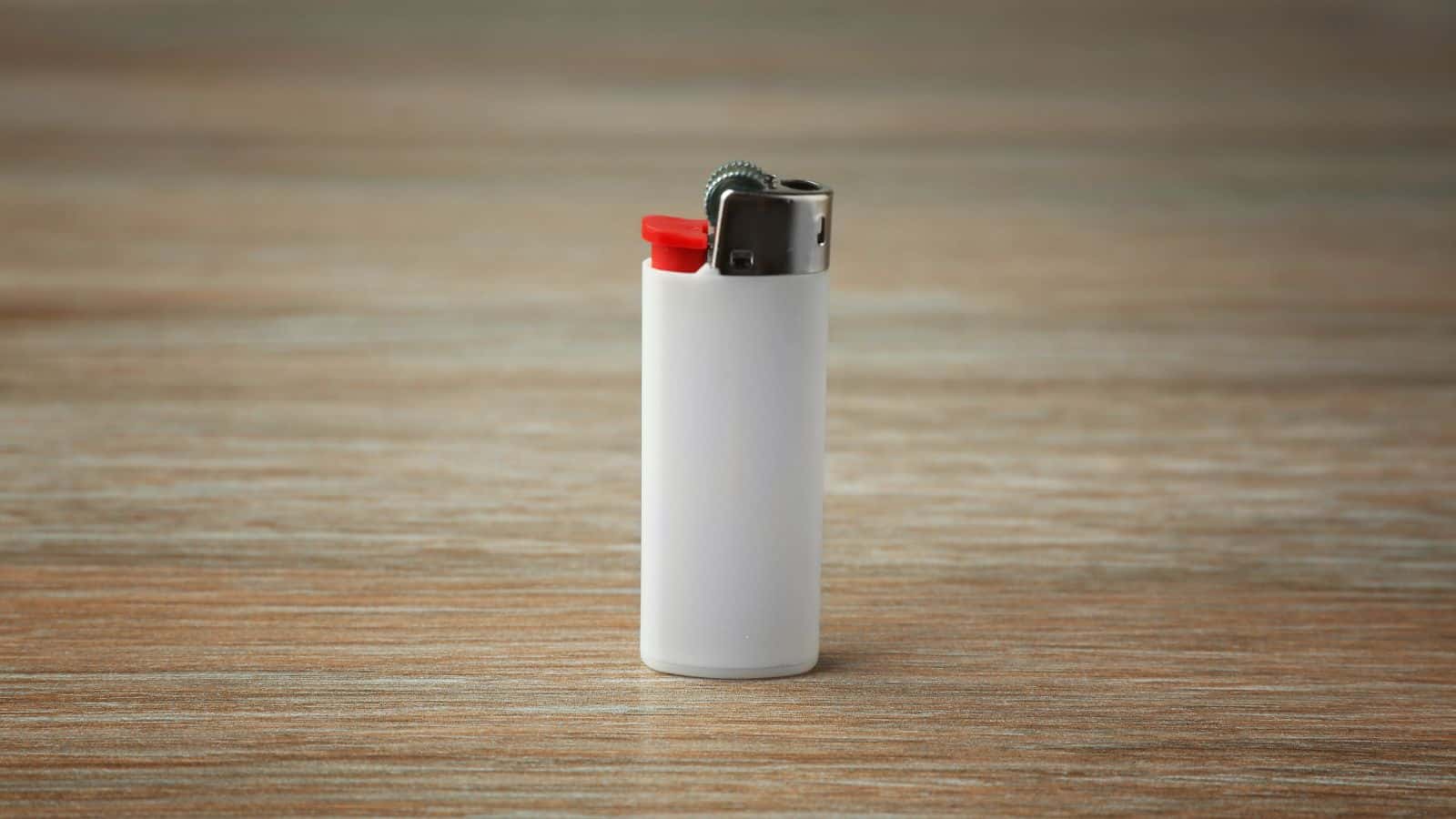 A white disposable lighter with a red ignition button is standing upright on a wooden surface. The metal top is slightly tarnished, and the background is softly blurred.