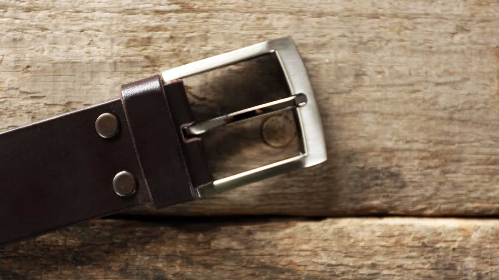 A close-up of a brown leather belt with a silver buckle resting on a wooden surface. The belt has two metal rivets near the buckle. The wood has a rough, textured appearance with visible grains.