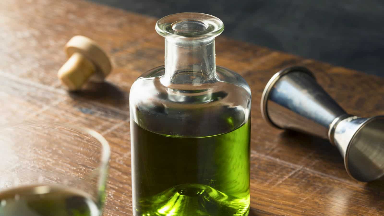 A clear glass bottle filled with green liquid sits on a wooden surface. The bottle is sealed with a cork stopper. A metal jigger lies on its side nearby, and part of a glass is visible in the foreground.