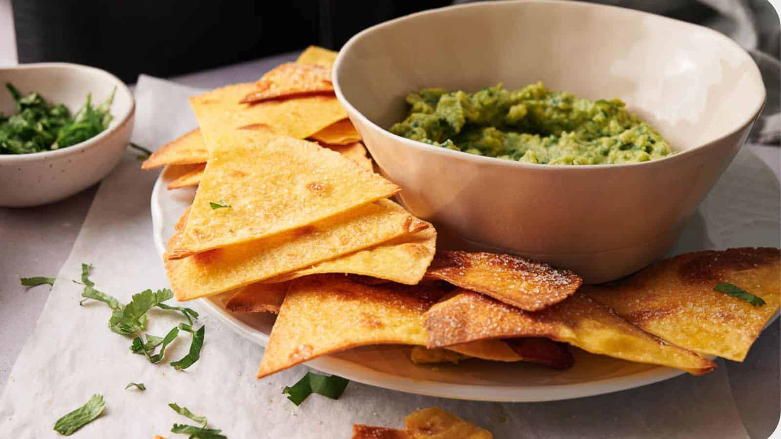 A plate of crispy tortilla chips is arranged next to a bowl of chunky guacamole. There is a small bowl with chopped herbs in the background, placed on a table covered with parchment paper.