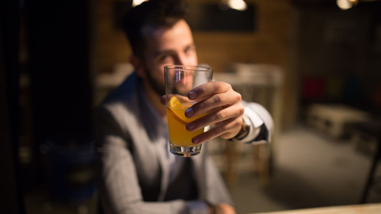 A person in a suit holds a glass with an orange liquid and an orange slice, extending it towards the camera in a dimly lit setting. The background is blurred, featuring a table and chairs.