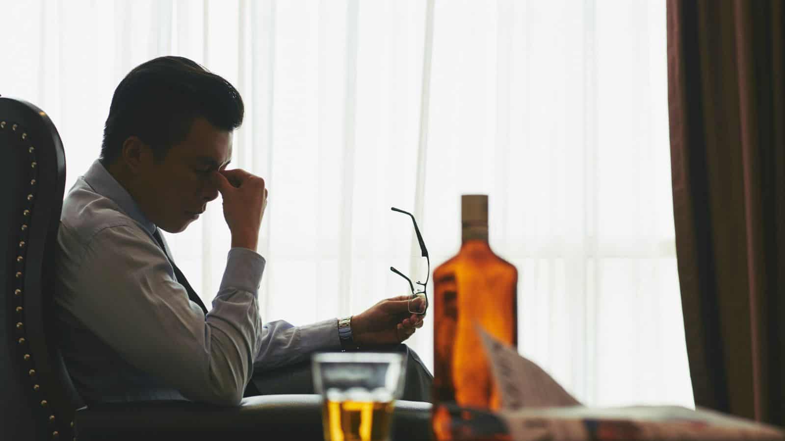 A man sits on a chair in a dimly lit room, rubbing his forehead while holding a pair of glasses. A bottle and glass of amber liquid are on a nearby table. He appears contemplative or stressed, with a window backlighting the scene.