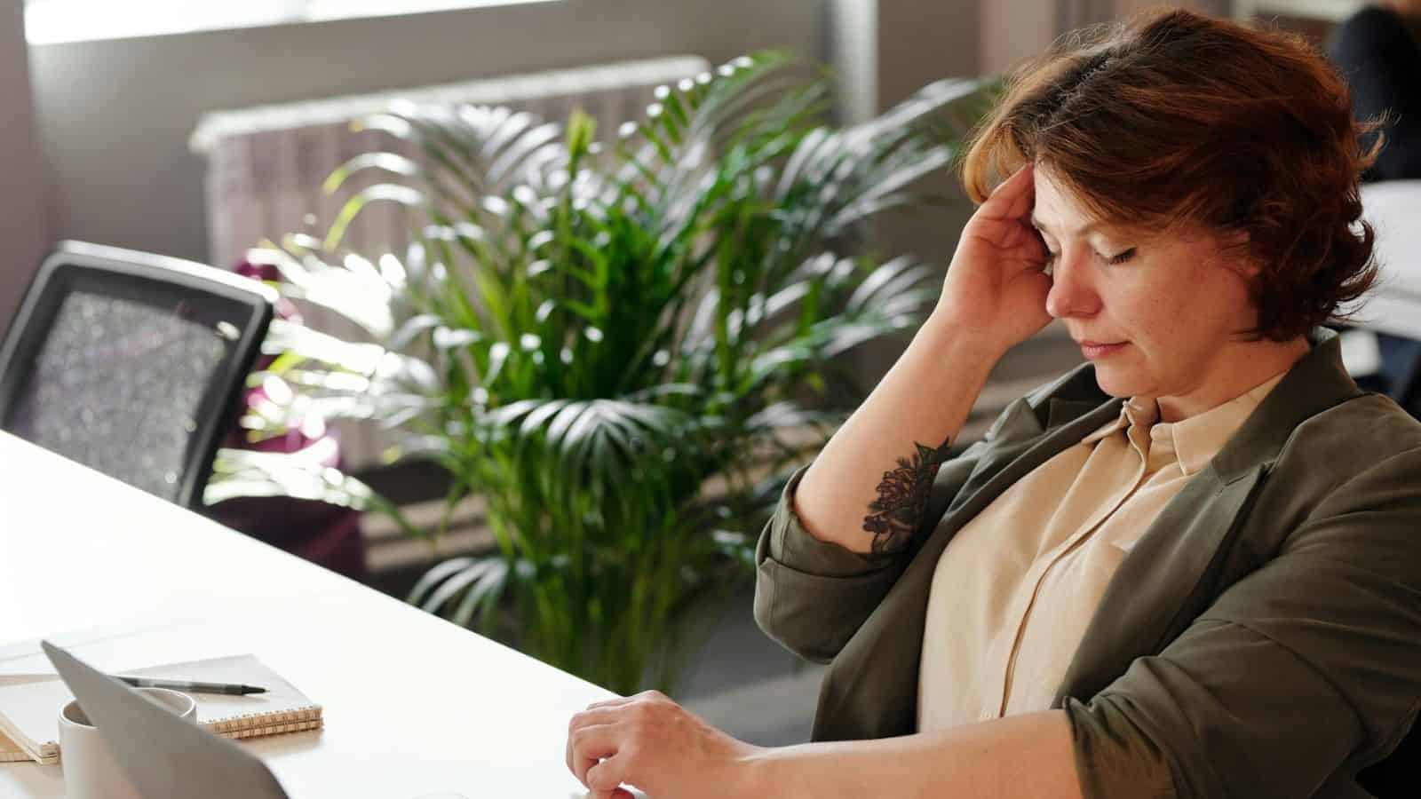 A person sitting at a desk with their hand on their forehead, appearing thoughtful or stressed. There is a notebook and pen nearby, and a large plant in the background, with soft natural light coming in through a window.