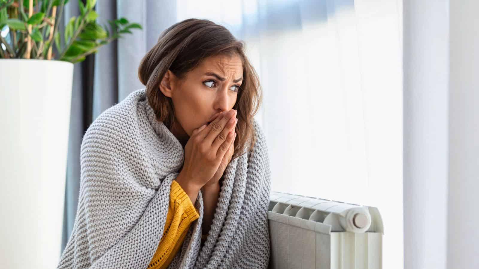 A woman wrapped in a blanket sits near a radiator, looking to the side with a concerned expression. She holds her hands close to her face, appearing to warm them. A potted plant is in the background.