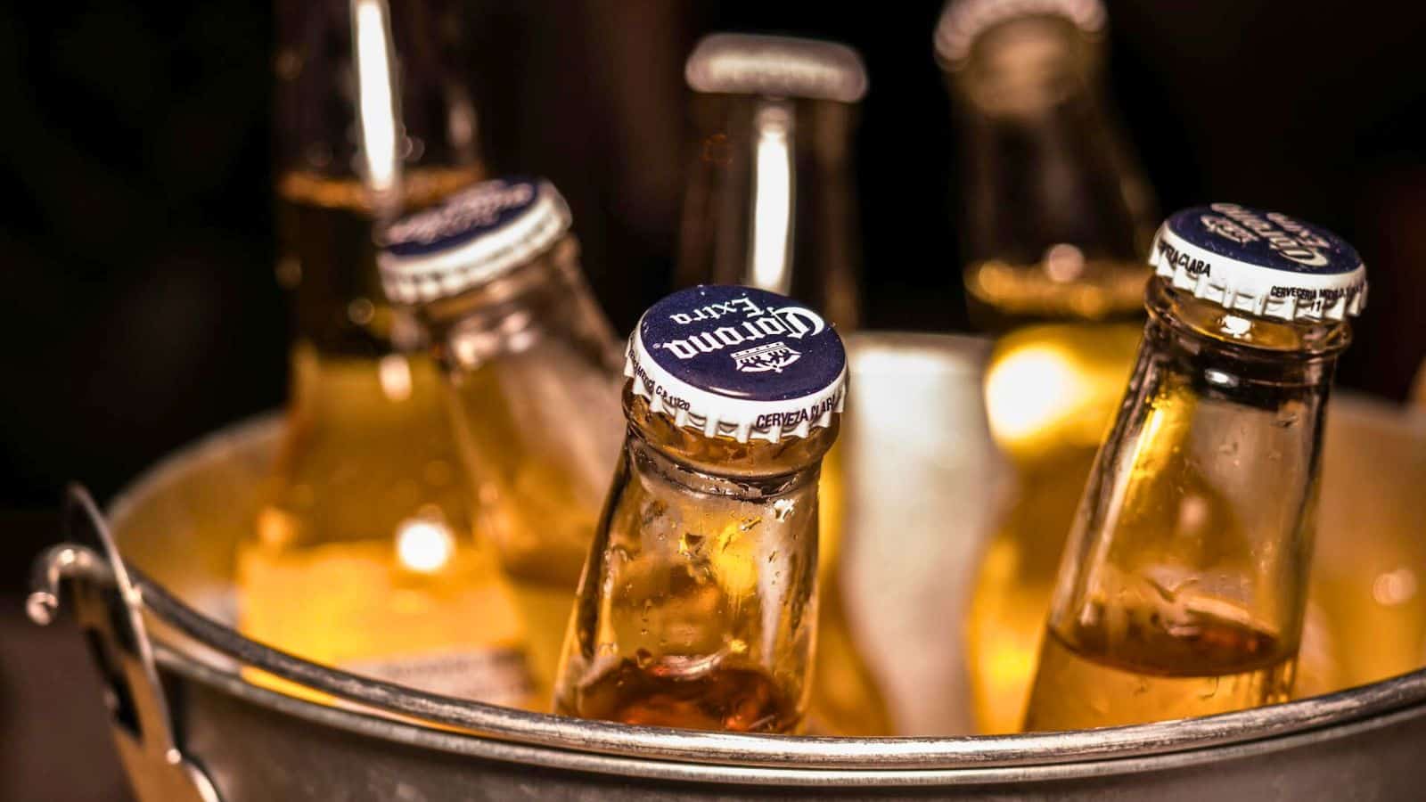 A metal bucket filled with several Corona beer bottles. The bottles are angled outward, with condensation visible on the glass. The blue and white caps are prominent, and the ambient lighting gives a warm hue to the scene.