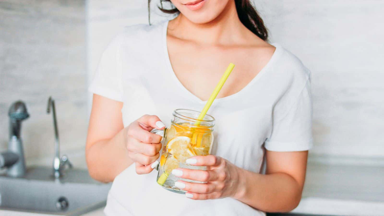 A person in a white shirt holds a glass mason jar filled with a lemon-infused drink and a yellow straw. The background features a kitchen sink and light-colored walls.