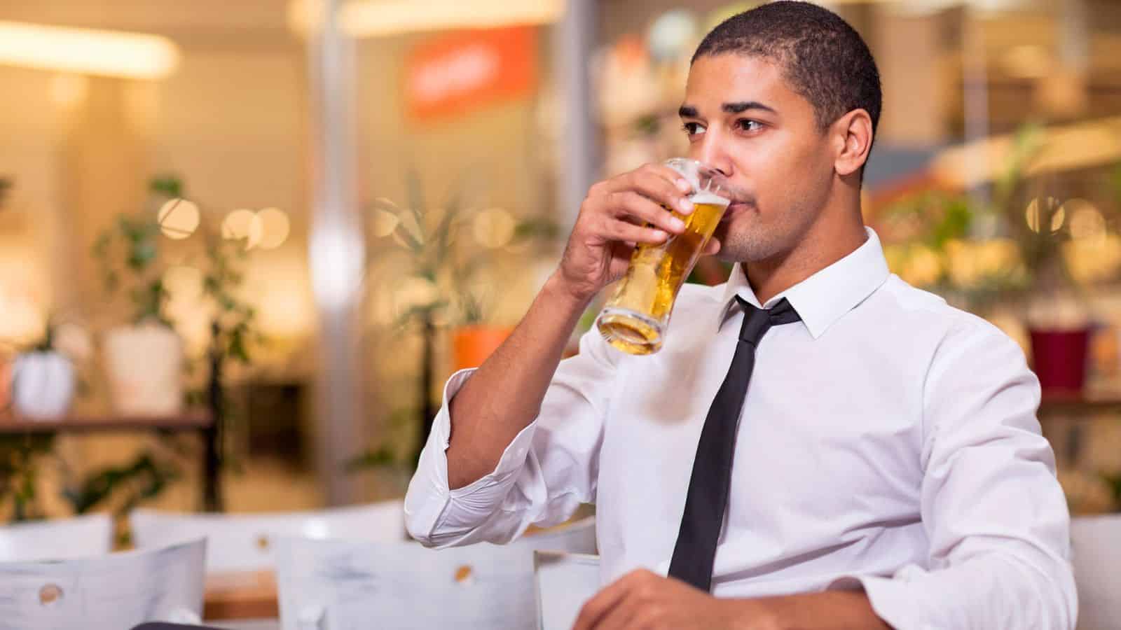 A man in a white shirt and black tie is sitting at a table in a bright indoor space, drinking from a glass of beer. There are blurred figures and plants in the background.