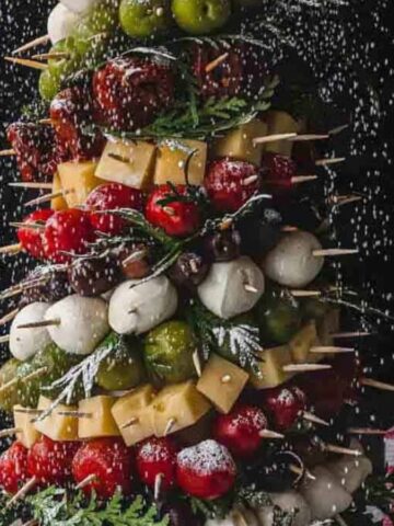 A festive skewer tree with layers of assorted cheeses, tomatoes, olives, and cured meats, topped with a tomato and garnished with herbs. Perfect for holiday gatherings, this centerpiece pairs beautifully with festive appetizers and drinks. A jar of olives and gifts complete the scene.
