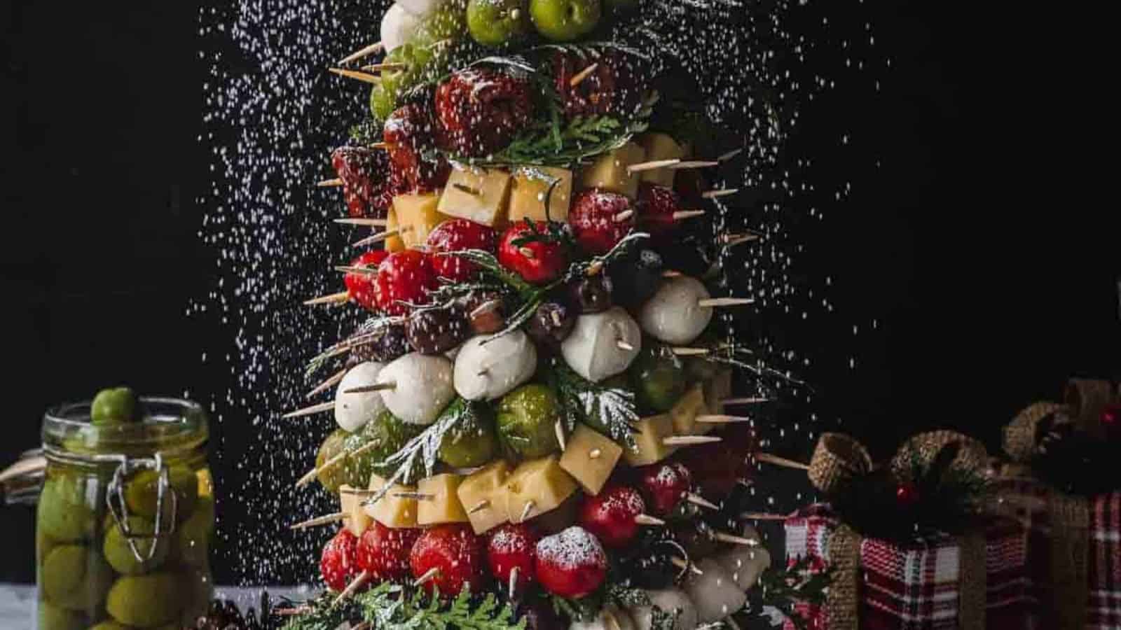 A festive skewer tree with layers of assorted cheeses, tomatoes, olives, and cured meats, topped with a tomato and garnished with herbs. Perfect for holiday gatherings, this centerpiece pairs beautifully with festive appetizers and drinks. A jar of olives and gifts complete the scene.