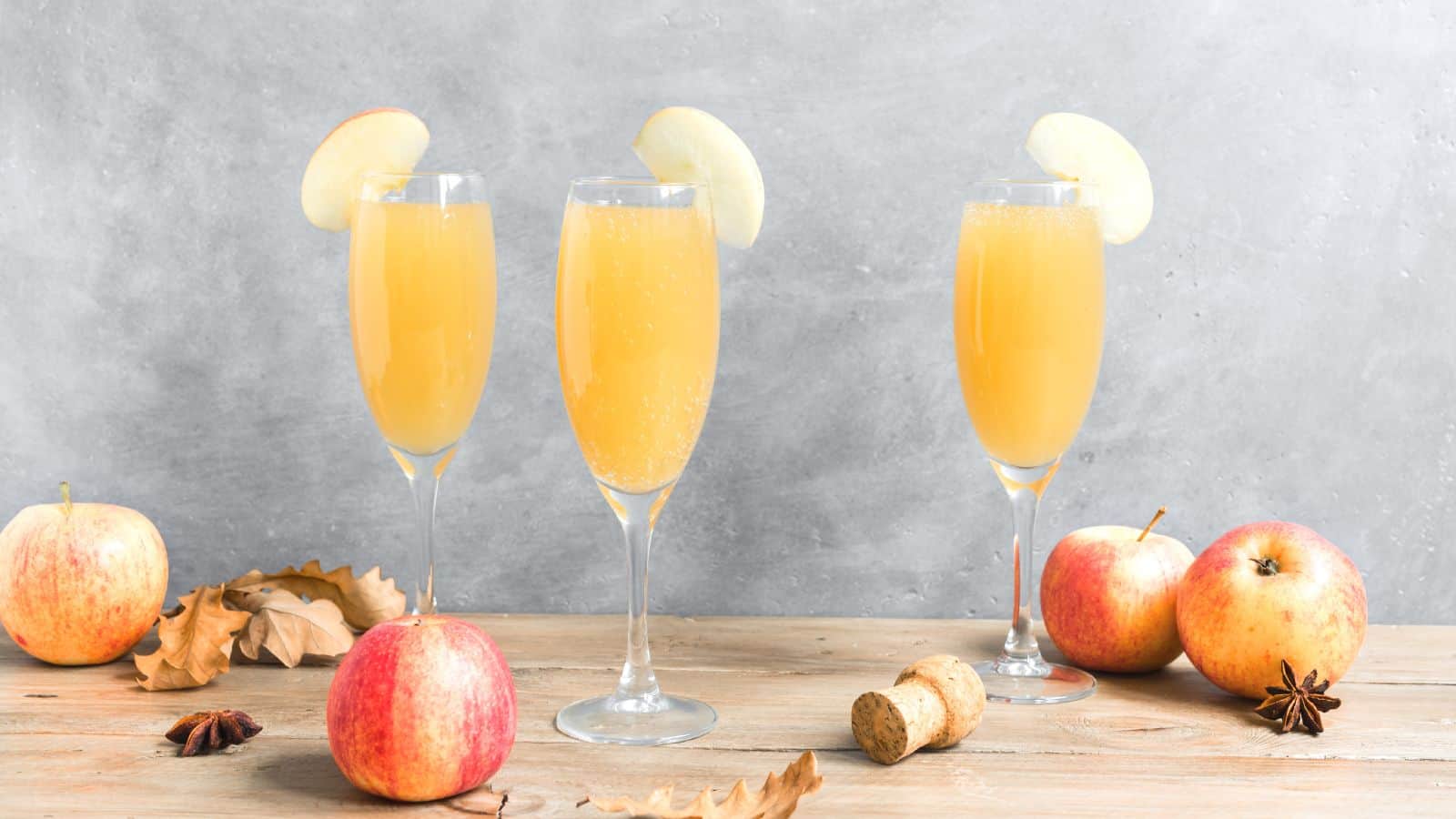 Three champagne flutes filled with apple juice and garnished with apple slices are on a wooden surface. Surrounding the glasses are whole apples, a cork, star anise, and dried leaves, against a gray background.