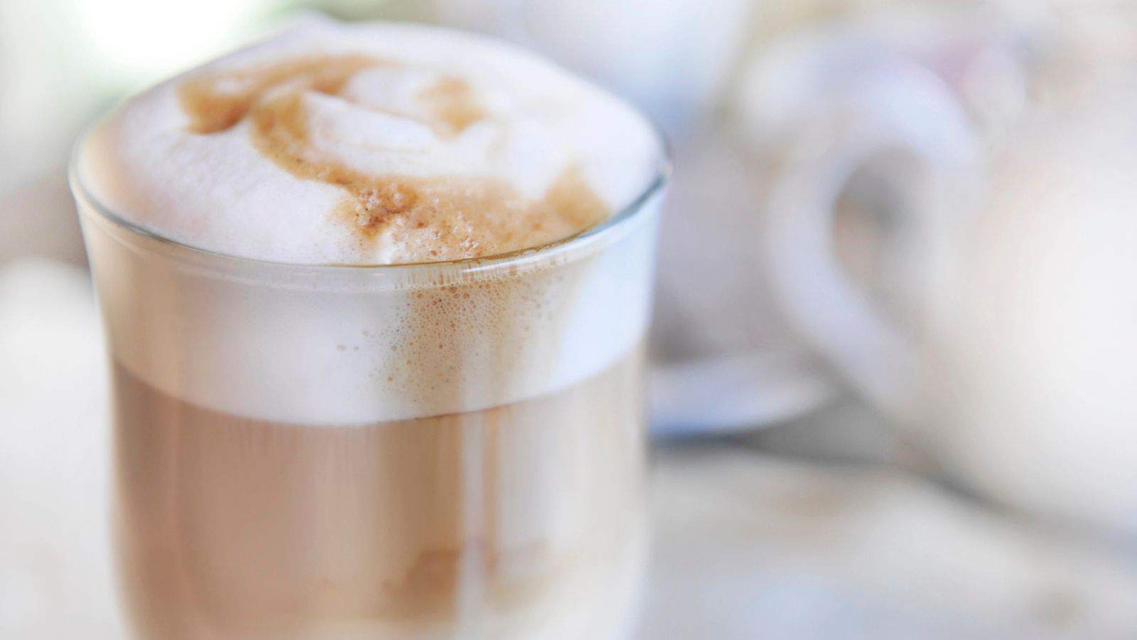 A glass cup of cappuccino with frothy milk and a hint of caramel drizzle on top. The background is blurred, featuring subtle light tones.