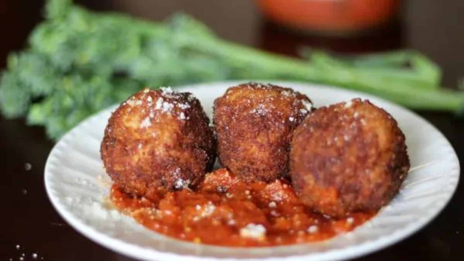 Three fried arancini balls sit on a white plate over a spread of tomato sauce, garnished with grated cheese. In the background, there is a branch of broccoli.