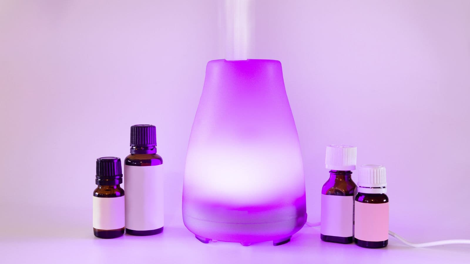 A purple essential oil diffuser emits mist, surrounded by five small bottles of essential oils with labels in various colors. The setup is against a light pink background.