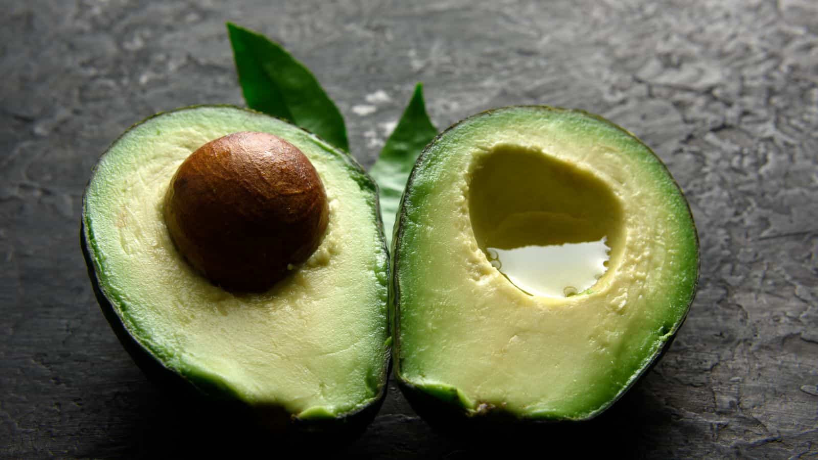 A halved avocado is placed on a dark textured surface. One half contains the seed, while the other half is empty. There are two green leaves in the background.
