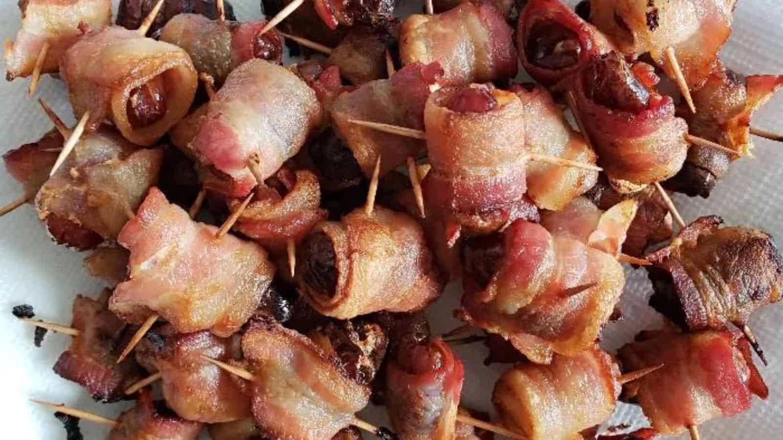 A platter of bacon-wrapped dates secured with toothpicks, arranged on a white paper towel. The bacon is crisp, and the dates have a dark, caramelized appearance.