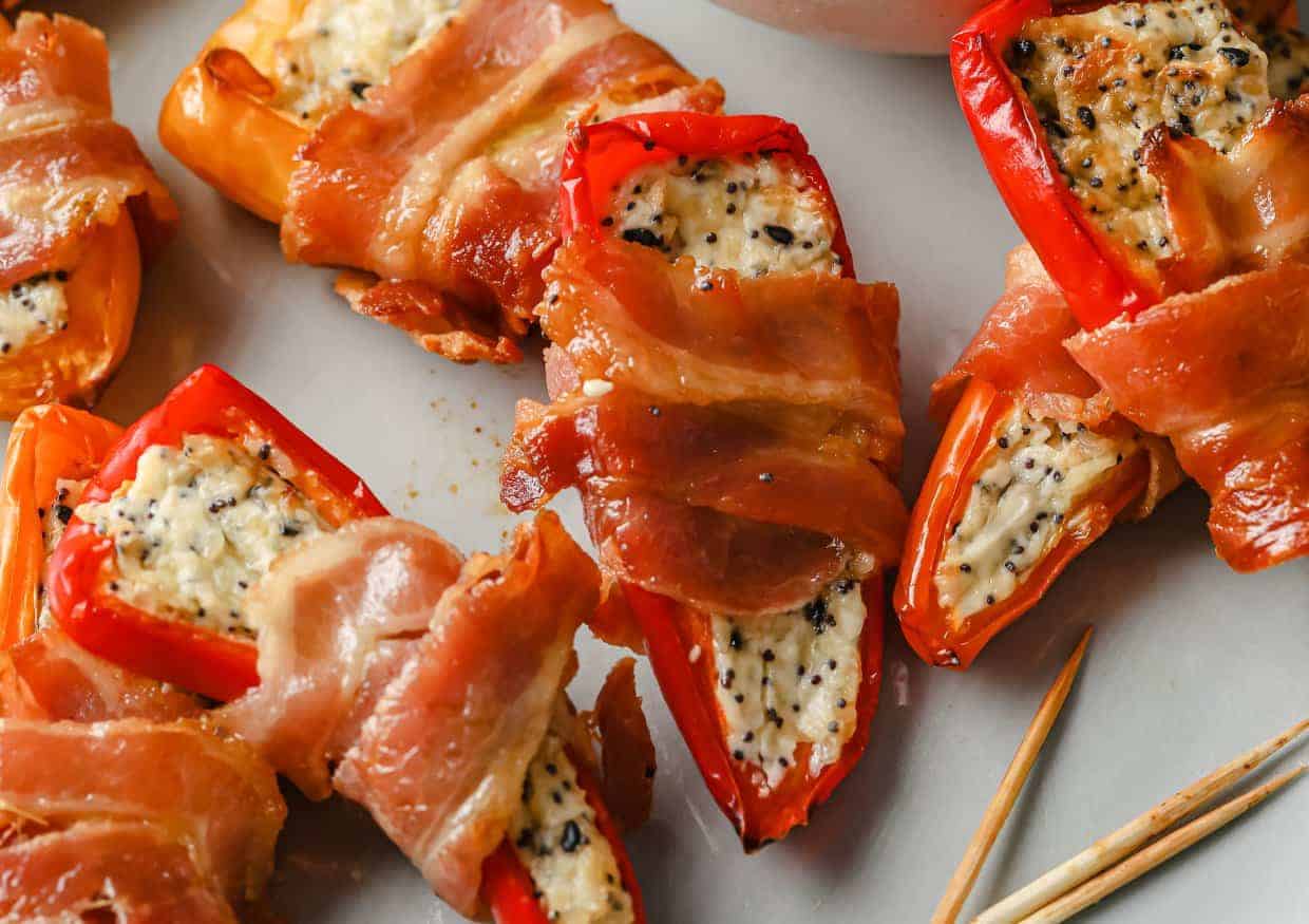 Bell peppers stuffed with a creamy filling and wrapped in crispy bacon are arranged on a plate. A few wooden toothpicks are visible beside them.