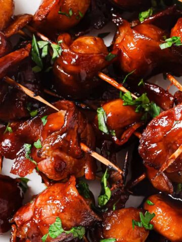 Close-up of bacon-wrapped water chestnuts on skewers, glazed with a sticky sauce and garnished with chopped parsley. The skewers are arranged on a white plate.