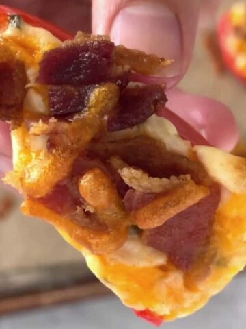 A hand holding a piece of stuffed bell pepper topped with cheese, bacon bits, and crispy onions. The background shows a baking tray with more stuffed pepper pieces.