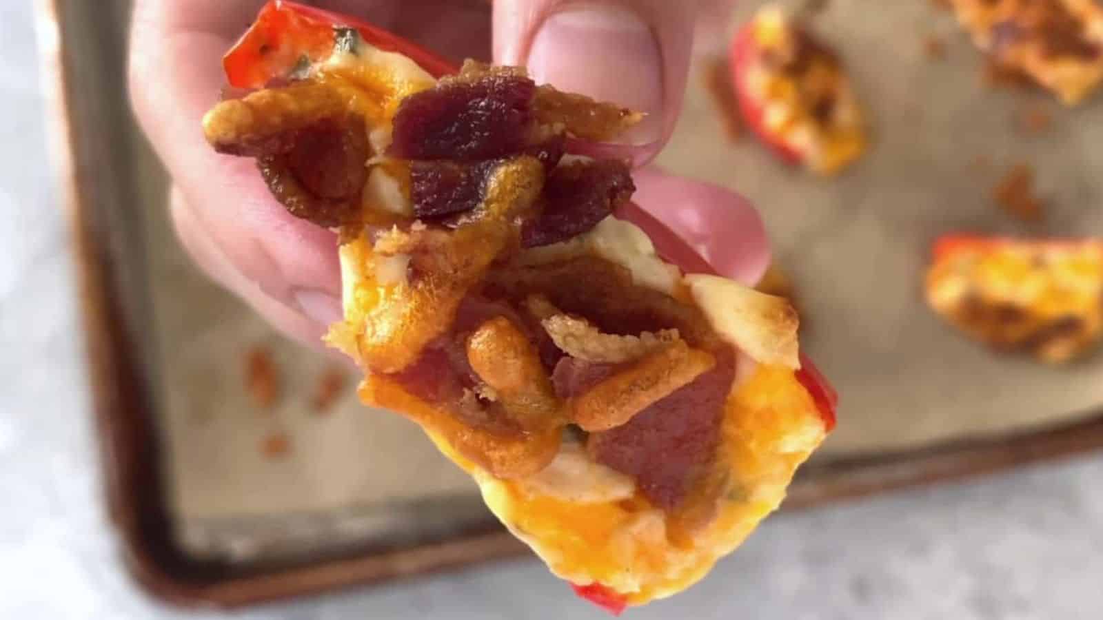 A hand holding a piece of stuffed bell pepper topped with cheese, bacon bits, and crispy onions. The background shows a baking tray with more stuffed pepper pieces.