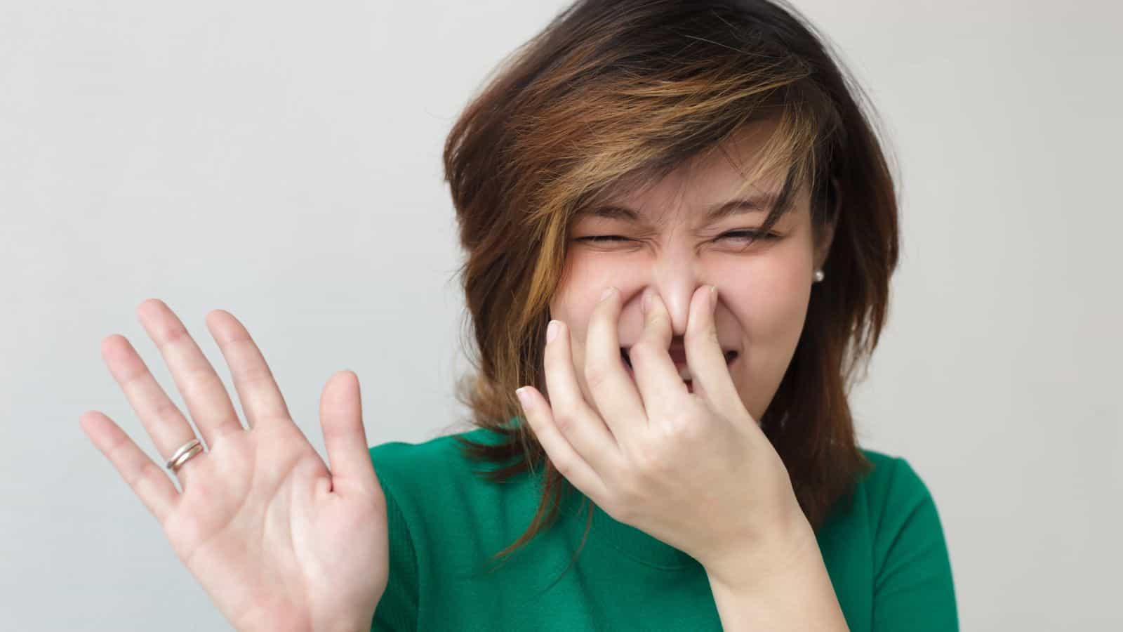 A person with medium-length hair is pinching their nose and has their eyes closed. They are wearing a green shirt and have one hand raised. The background is plain and light-colored.