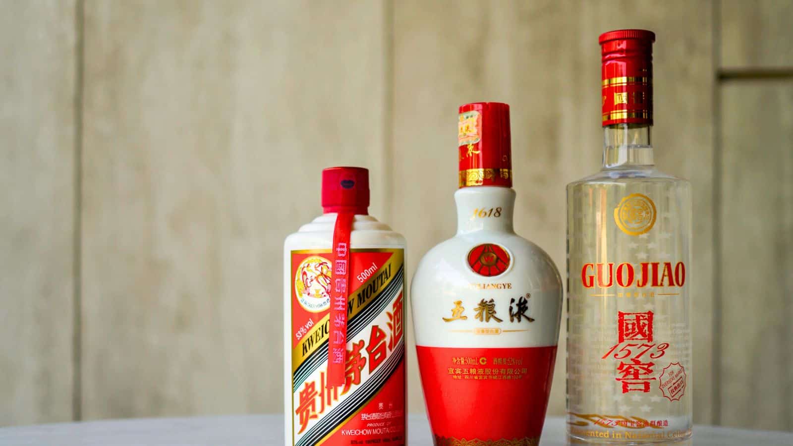 Three bottles of Chinese liquor are displayed on a surface. The labels feature red and gold designs, with distinct bottle shapes. The backdrop is a light beige wall.