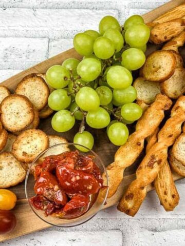 A wooden platter with grapes, a round bread bowl with melted cheese, sliced apples, cherry tomatoes, roasted red peppers in a glass, breadsticks, and toasted baguette slices, set against a white brick background.