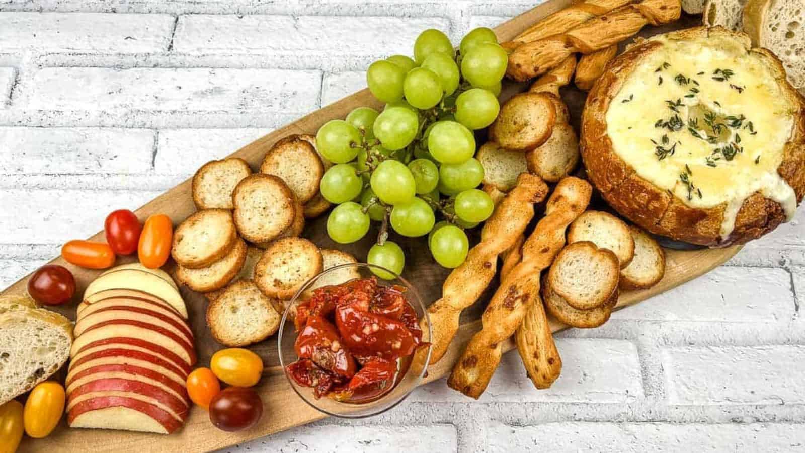 A wooden platter with grapes, a round bread bowl with melted cheese, sliced apples, cherry tomatoes, roasted red peppers in a glass, breadsticks, and toasted baguette slices, set against a white brick background.