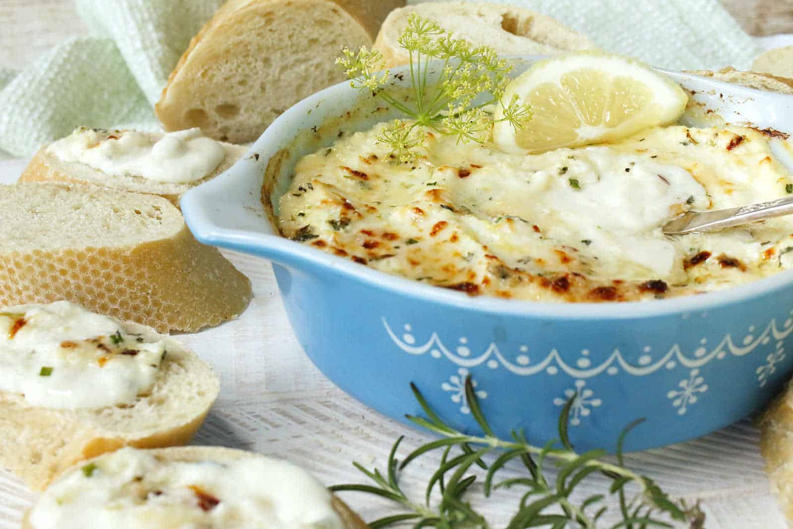 A blue ceramic dish contains a baked cheese dip with a lemon slice on top. Fresh herbs and slices of bread are arranged around the dish. A piece of bread is spread with the dip.