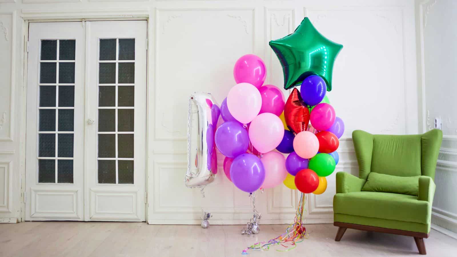 A cluster of colorful balloons including pink, purple, green, and star-shaped, is tied to a silver weight in a room with white paneled walls. A light green armchair is positioned nearby, and double glass doors are in the background.